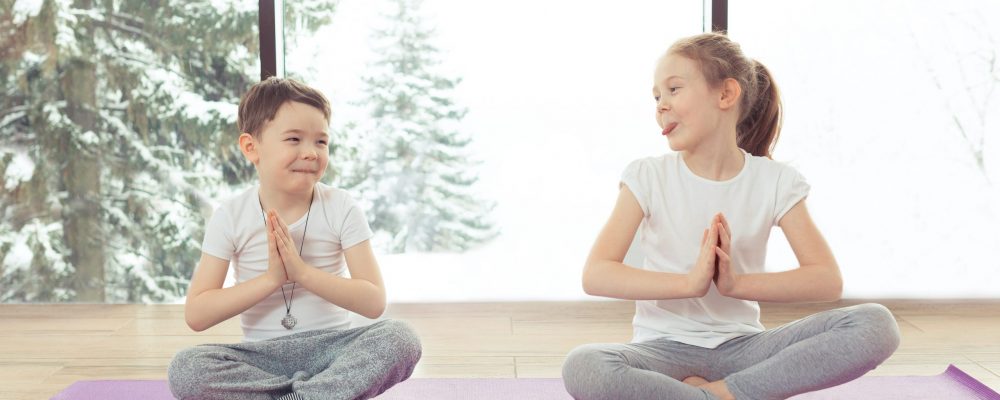 Group of children doing gymnastic yoga exercises at the winter resort with a beautiful view from the window. Two kids brother and sister doing lotus pose and smiling together