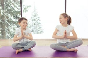 Group of children doing gymnastic yoga exercises at the winter resort with a beautiful view from the window. Two kids brother and sister doing lotus pose and smiling together